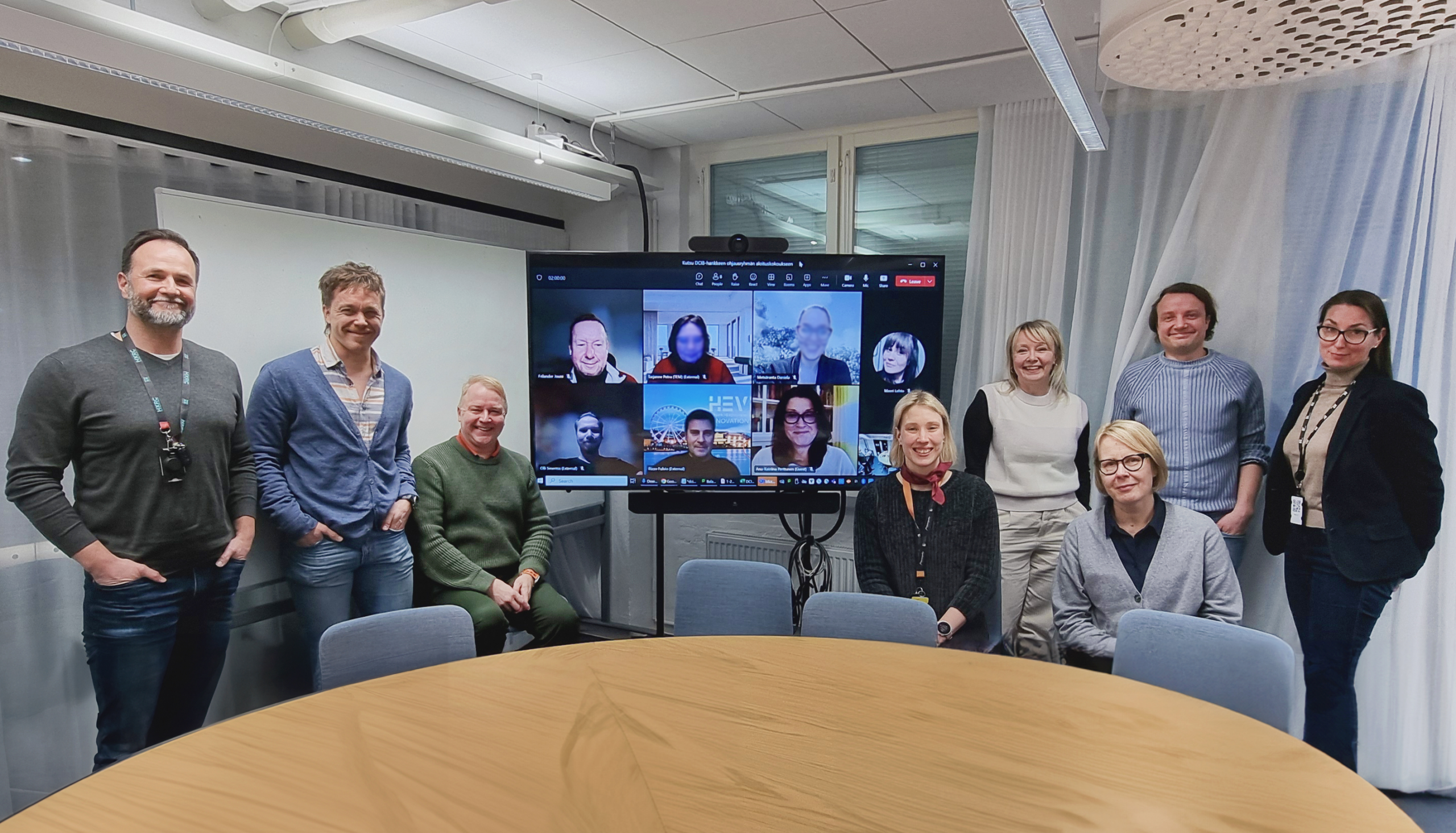 DCIB-hankkeen ohjausryhmä yhteiskuvassa ensimmäisen ohjausryhmän tapaamisen päätteeksi pyöreän pöydän ääressä tammikuussa 2024. Kahdeksan henkeä on fyysisesti tilassa, ja seitsemän henkeä näkyvät etätyöpisteiltään televisioruudun välityksellä.