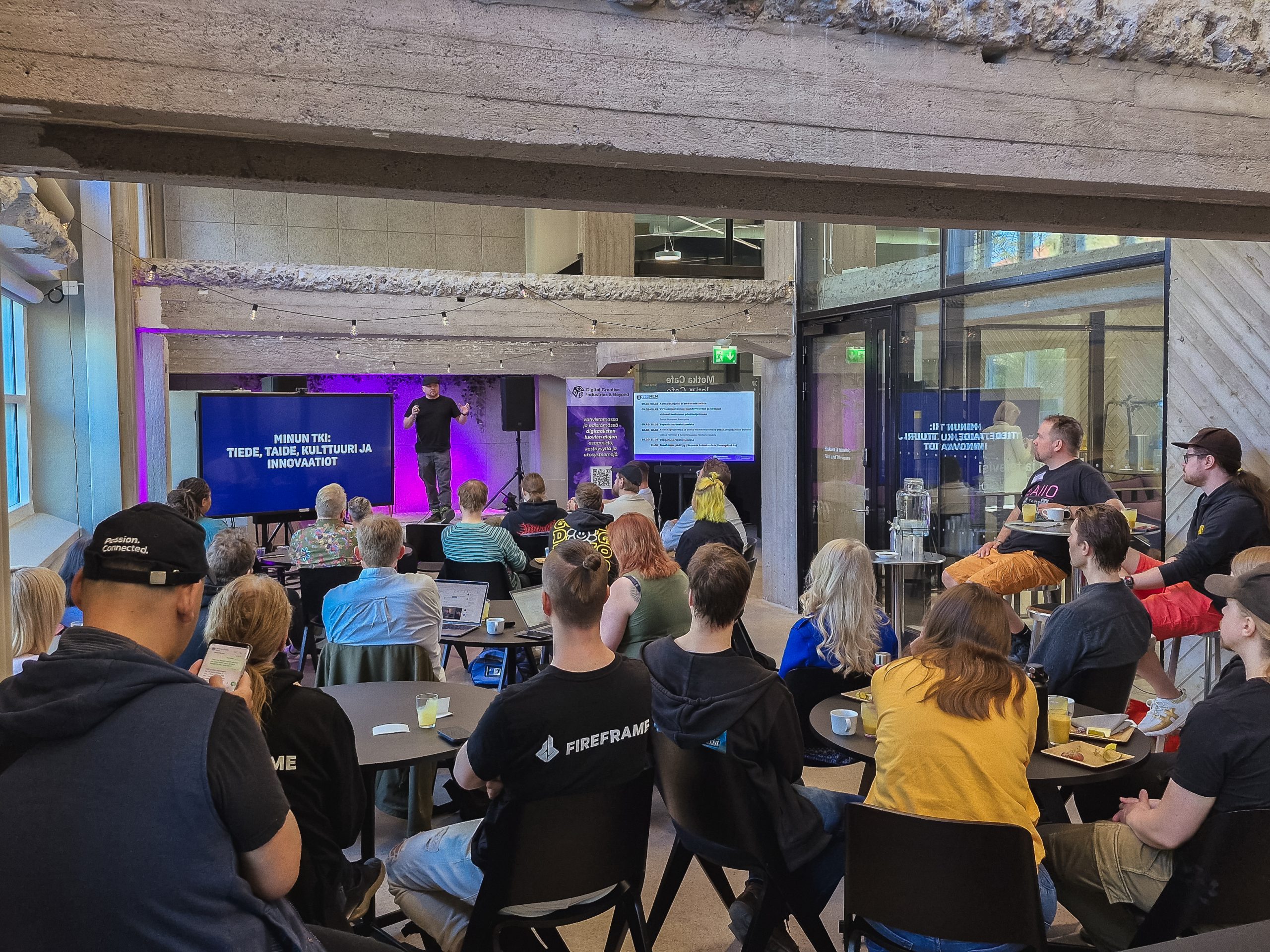 Samuli Homanen esiintymässä virtuaalituotantoteemaisessa heTKInen-aamukahvitapahtumassa 16. toukokuuta 2024. Yleisössä on muutama kymmenen ihmistä kerääntyneenä pöytäryhmien ääreen.