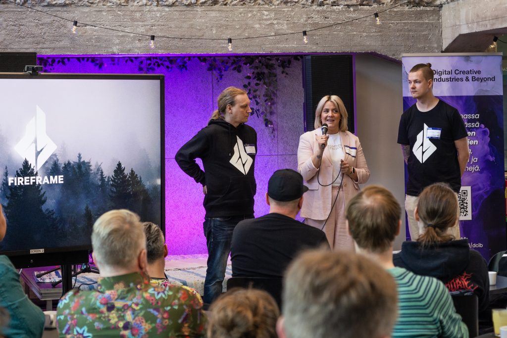 Fireframe Studiosin Markus Helminen, Annemi Kuusela ja Kasper Leppänen esiintymässä virtuaalituotantoteemaisessa heTKInen-aamukahvitapahtumassa 16. toukokuuta 2024. Yleisössä on muutama kymmenen ihmistä katsomasas heitä, kerääntyneenä pöytäryhmien ääreen.