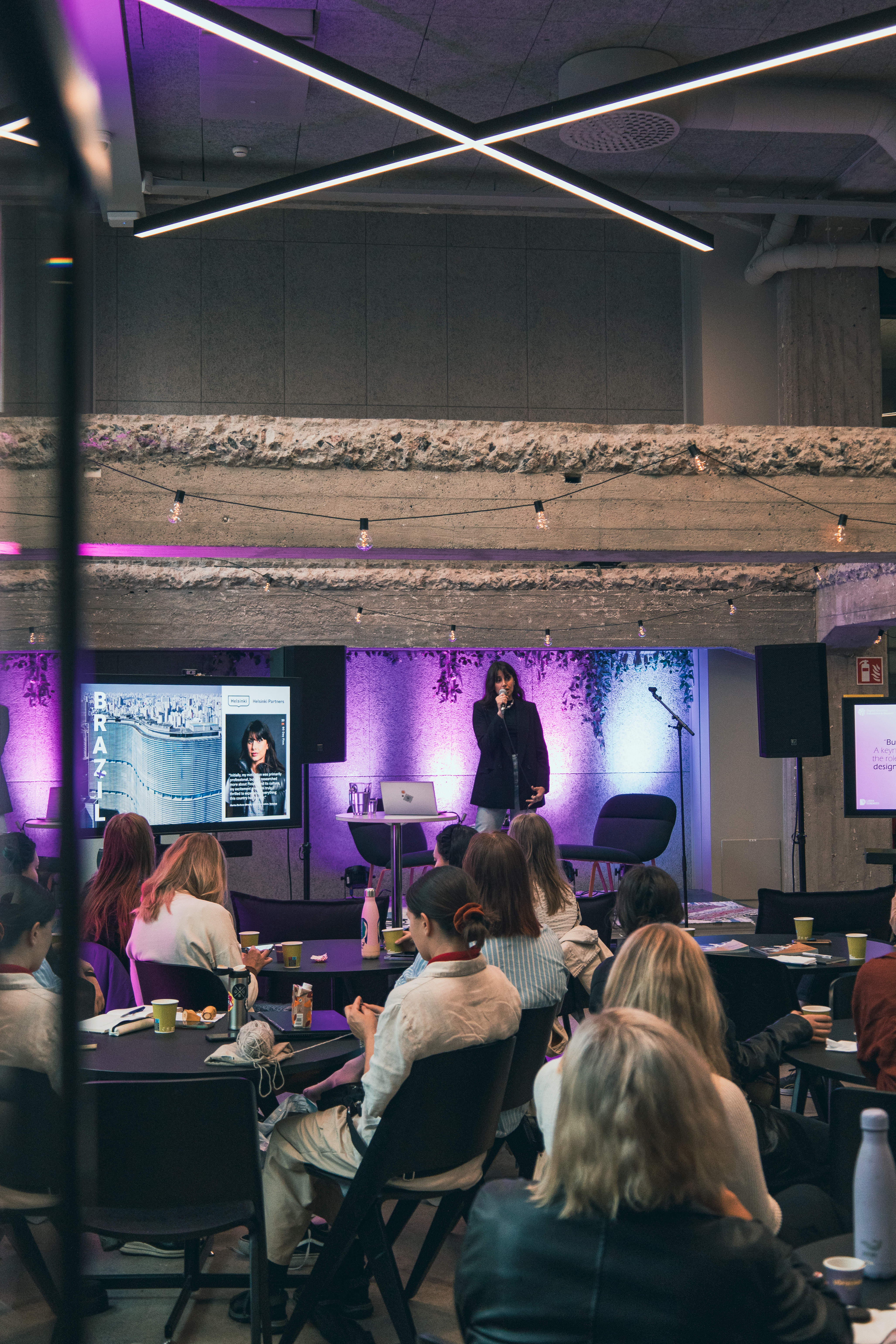 Marina Barboza standing on Monttu-stage in Metka Cafe, talking to the audience. The crowd is sitting and watching Marina speak. There's purple and cyan lights, making the high ceiling space look very cozy.