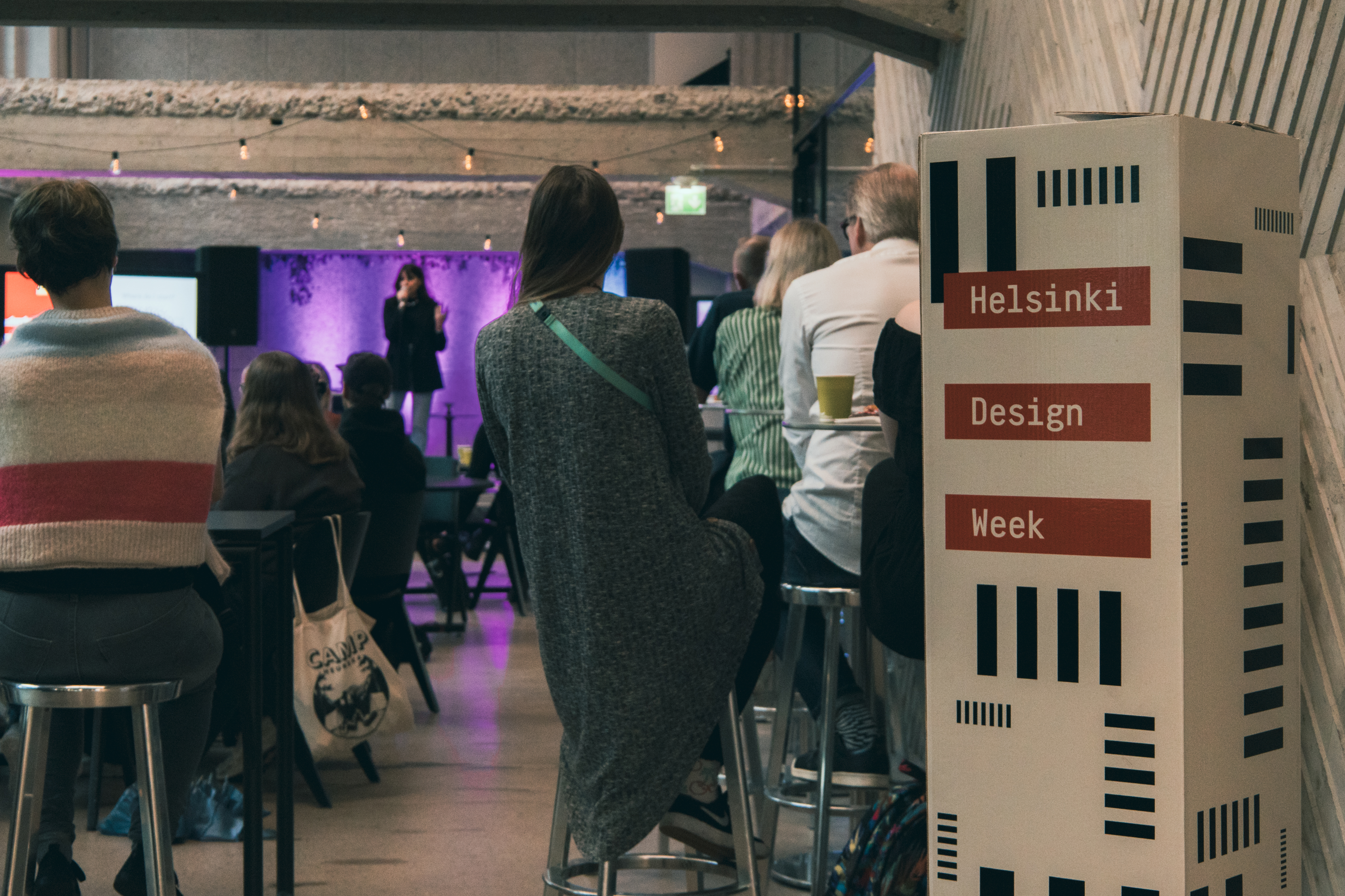 In the front is a cardboard box saying "Helsinki Design Week", indicating that this is a Helsinki Design Week. In the back, quite blurry Marina Barboza is standing on Monttu-stage in Metka Cafe, talking to the audience. The crowd is sitting and watching Marina speak. There's purple and cyan lights in the background.