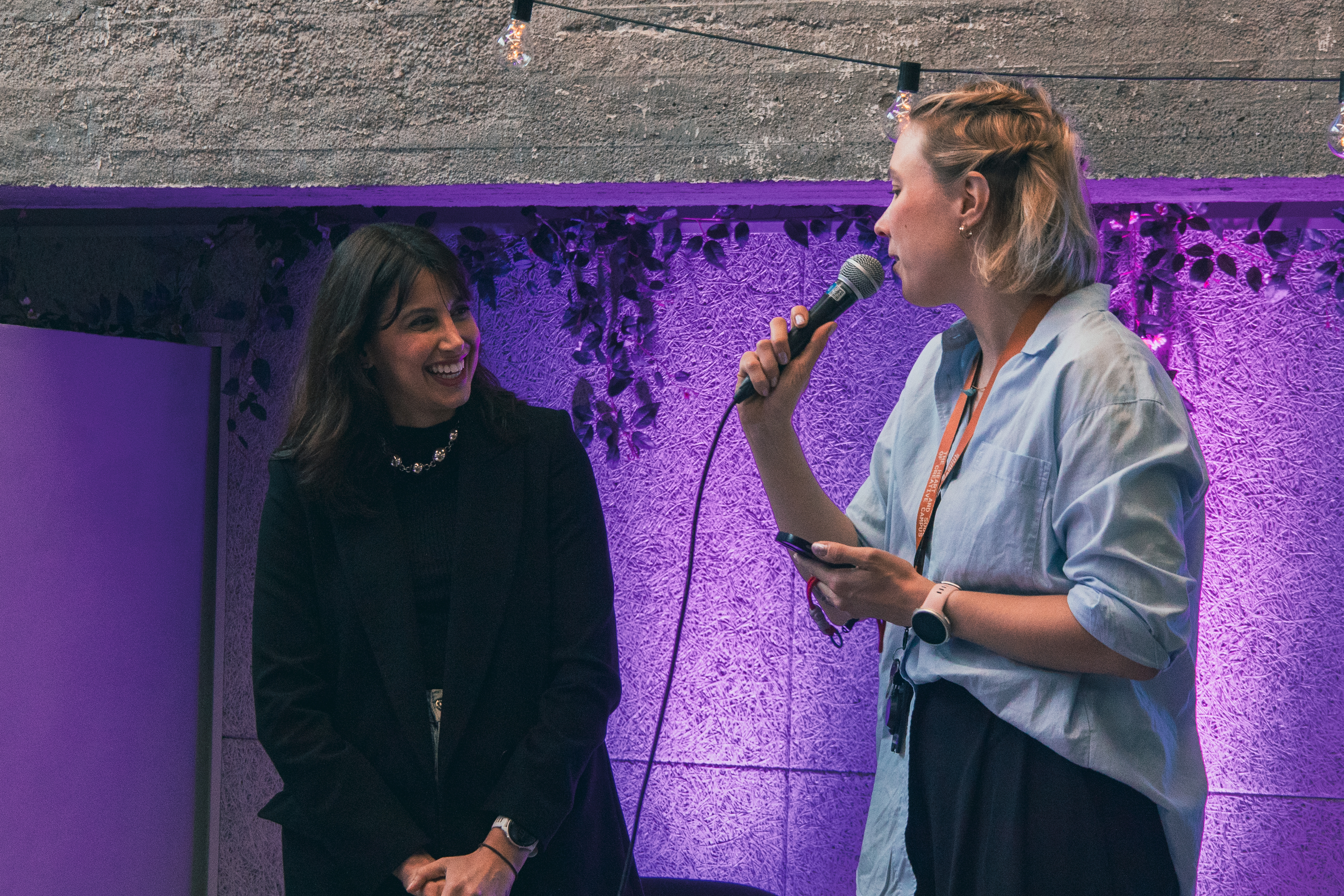 Marina Barboza standing on Monttu-stage in Metka Cafe, talking to Anna Muukkonen, who is Metropolia UAS project manager in the Digital Creative Industries and Beyond -project. Anna is holding the mic and asking Marina something, and Marina is laughing.