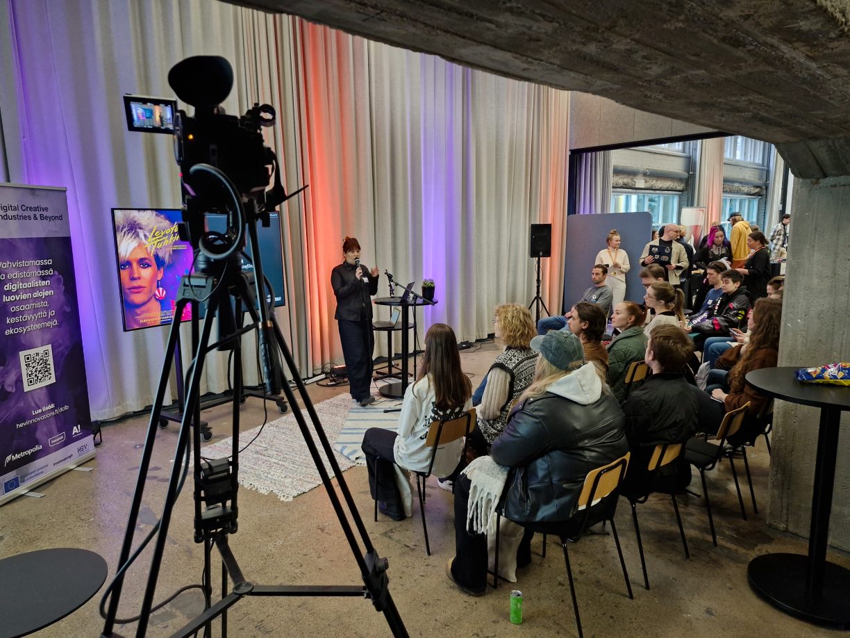 Yellow Film & TV esittäytymässä DCIB-hankkeen Digitaalisten luovien alojen työnantajatapahtumassa 27.2.2025. Etualalla näkyy livestream-kamera. Yleisö on täynnä porukkaa.