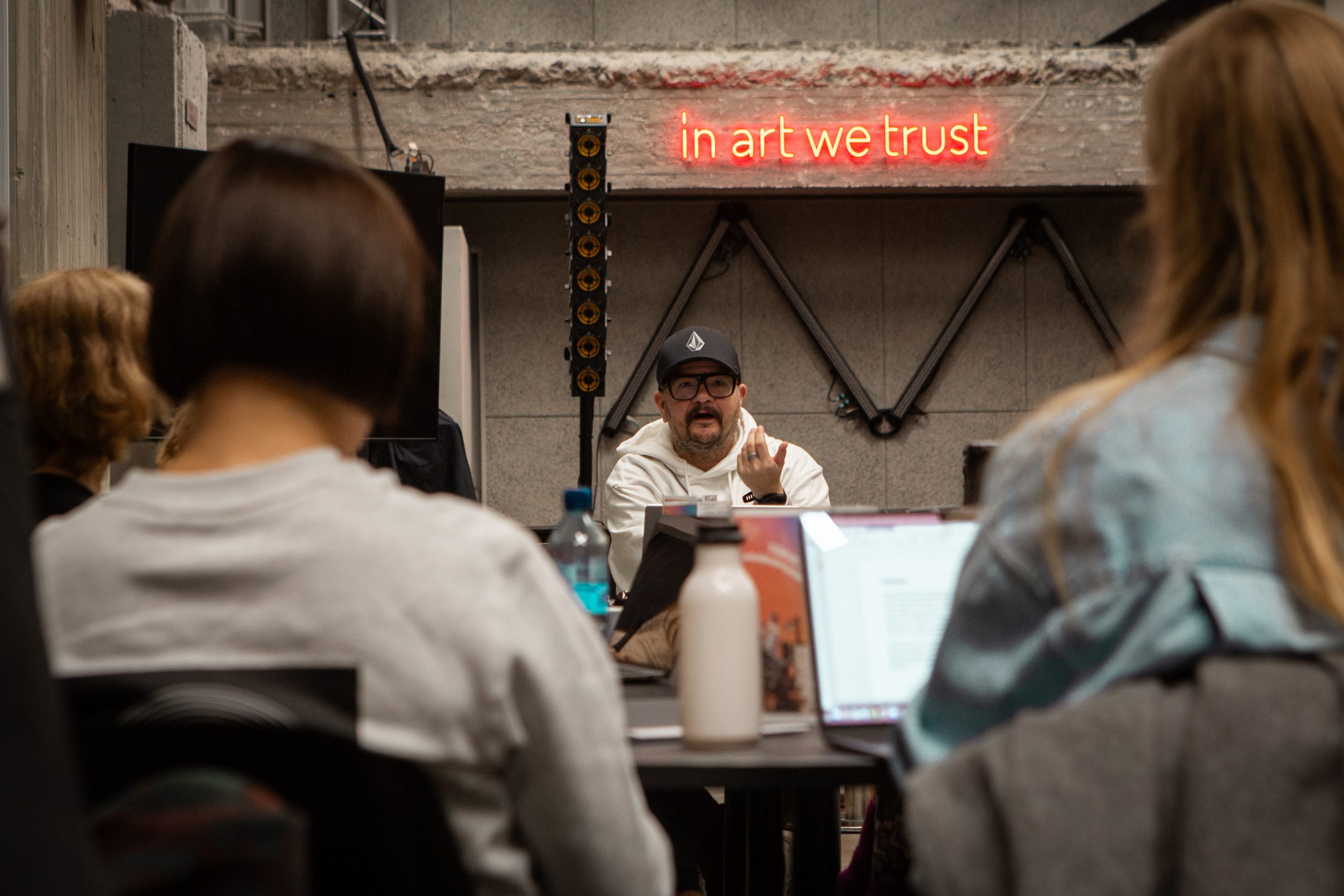 Ekokoordinoinnin pilottiohjelman vetäjä Taku Kaskela puhuu osallistujille. Hänen takanaan on neon kyltti, jossa lukee "in art we trust".